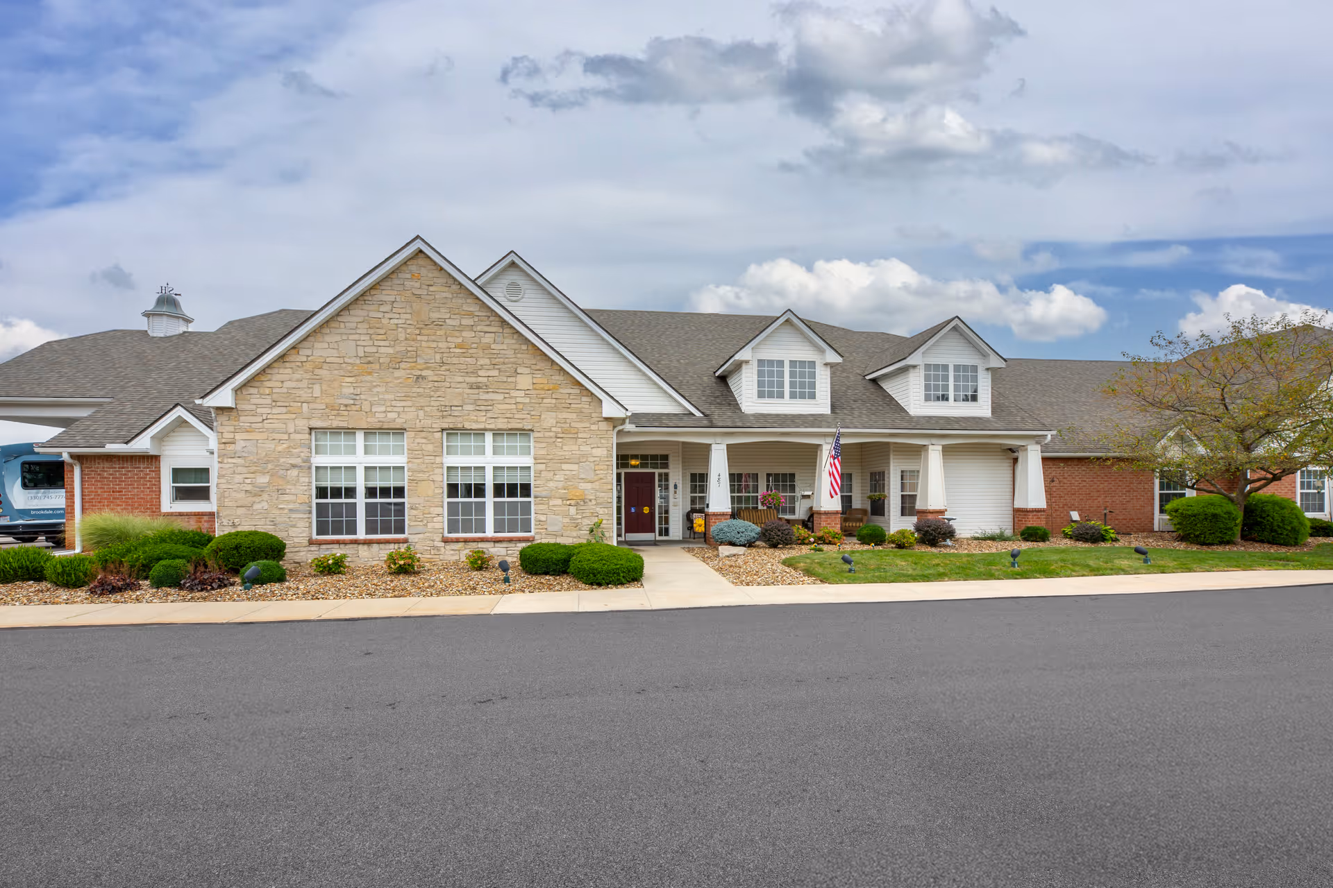 Front exterior view of Brookdale Barberton facility with a stone and brick facade, multiple windows, a covered entrance with an American flag, landscaped bushes, and a paved driveway.
