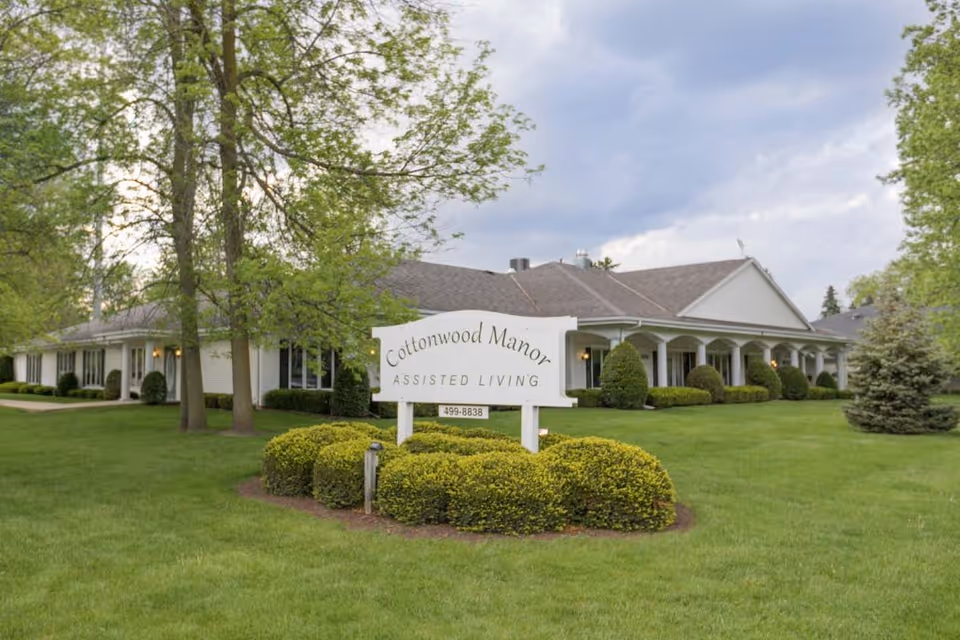 Exterior view of Cottonwood Manor Assisted Living facility showing a single-story building with a gray roof, white siding, and a covered porch with columns. The building is surrounded by green grass, bushes, and trees under a cloudy sky. A white sign in front reads 'Cottonwood Manor Assisted Living' with a phone number below.