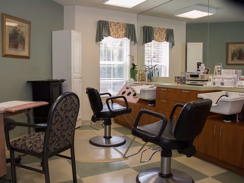 Interior of a salon room with two black salon chairs in front of sinks and wooden cabinets. There is a patterned armchair on the left side and two windows with floral valances letting in natural light. The walls are painted green and white, and there are framed pictures hanging on the walls.