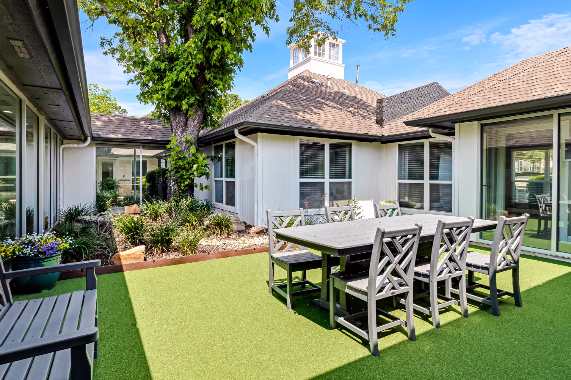 Outdoor courtyard area with a large tree surrounded by plants and rocks, a rectangular table with six chairs on artificial green turf, and windows of the surrounding building under a blue sky.