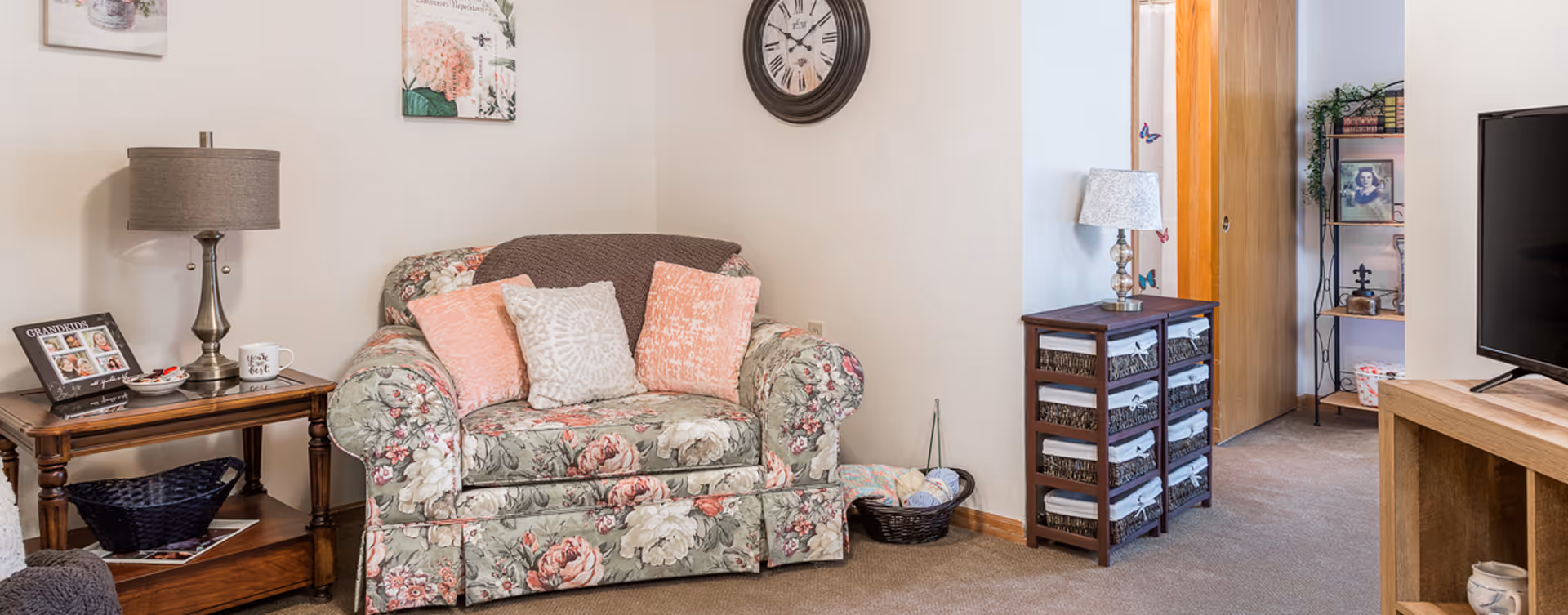 A cozy living room area with a floral patterned armchair adorned with three decorative pillows and a brown throw blanket. To the left is a wooden side table with a lamp, a photo frame labeled 'GRANDKIDS,' and a coffee mug. On the right side, there is a small wooden drawer unit with a lamp on top, a basket with yarn balls on the floor, and a metal shelf with books and framed photos. A large wall clock hangs above the armchair, and a flat-screen TV is partially visible on the right.