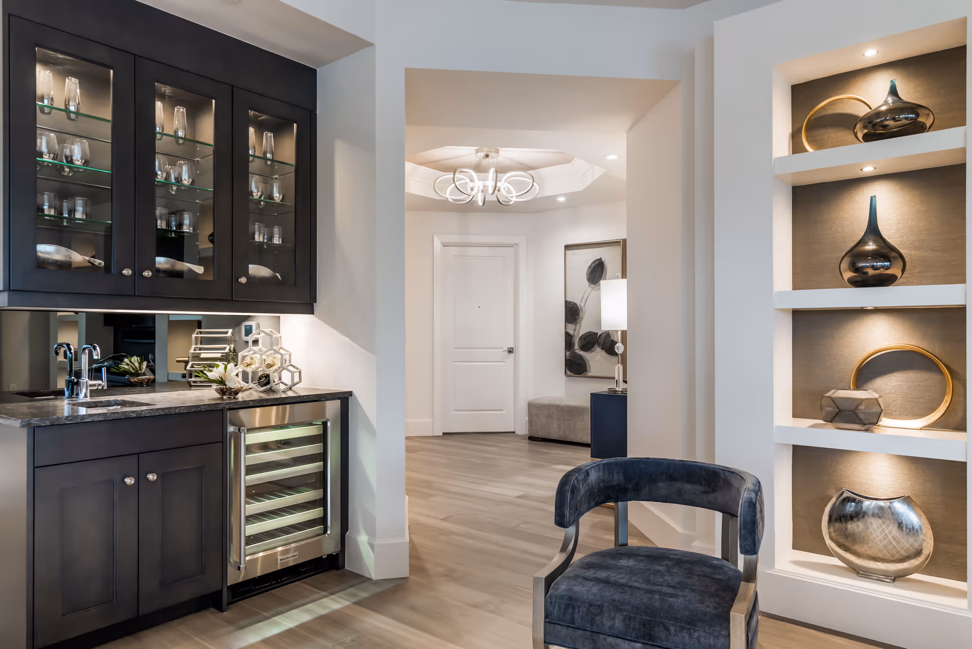 Bright modern interior showing a built-in wet bar with glass-front cabinets and wine fridge, decorative recessed shelving, a velvet chair, and an entry hallway.