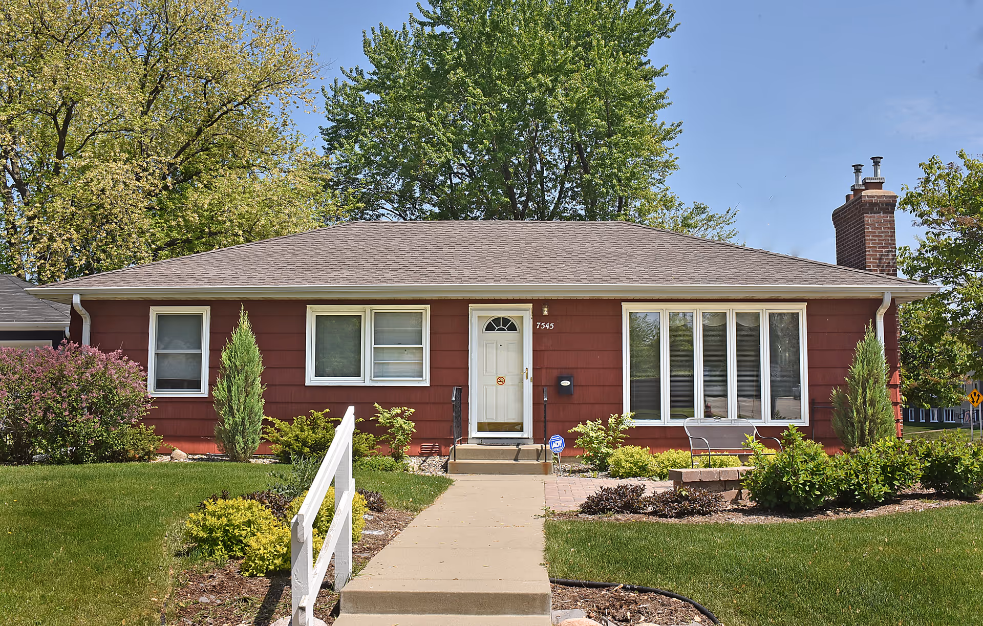 Single-story red house with a white front door and multiple windows, surrounded by green shrubs and trees under a clear blue sky. A concrete walkway with white railings leads to the front door.