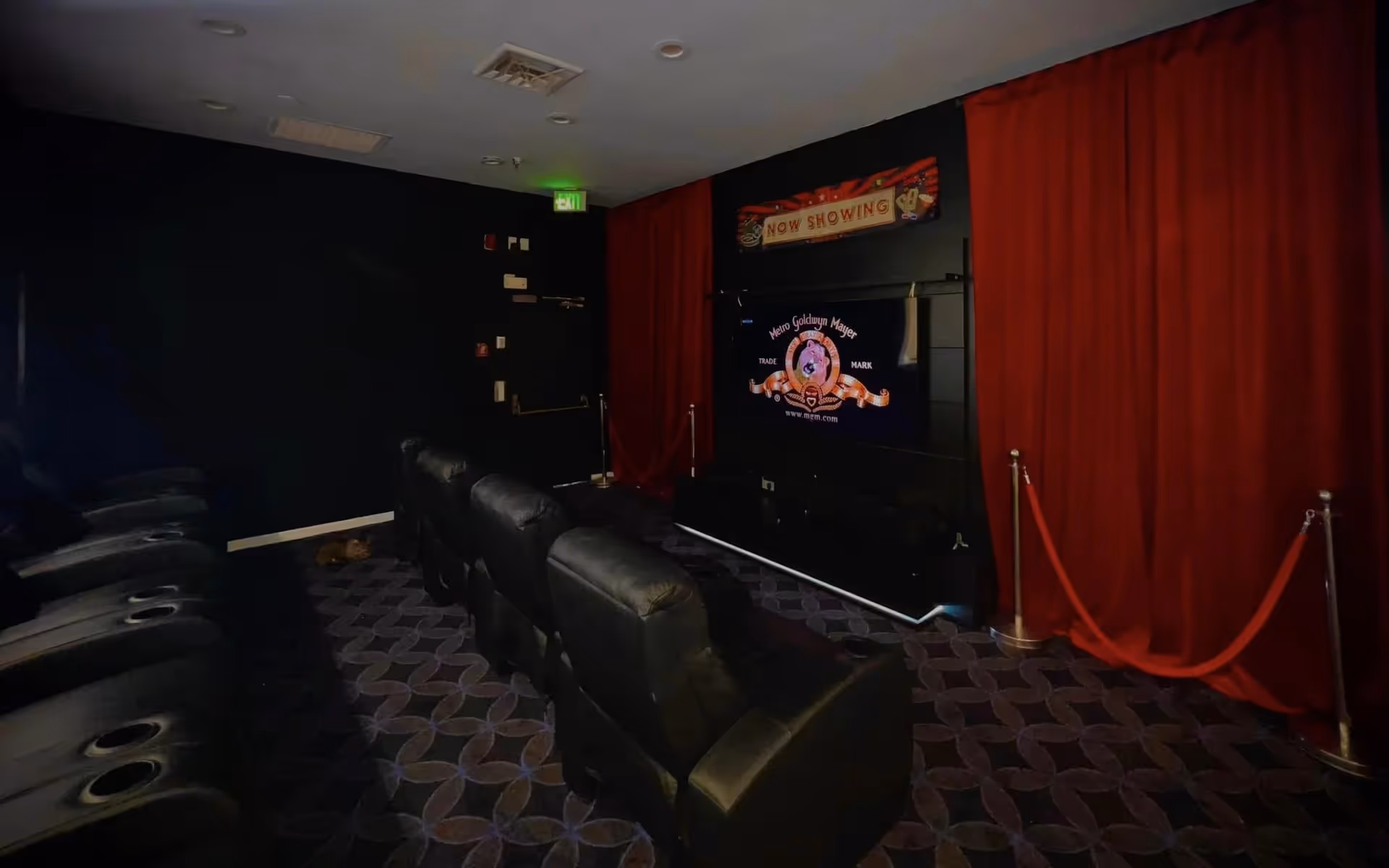 A small movie theater room with black leather recliner chairs arranged in rows facing a large screen displaying the Metro Goldwyn Mayer logo. The room has dark walls, a patterned carpet, red curtains on the right side, and a 'Now Showing' sign above the screen.