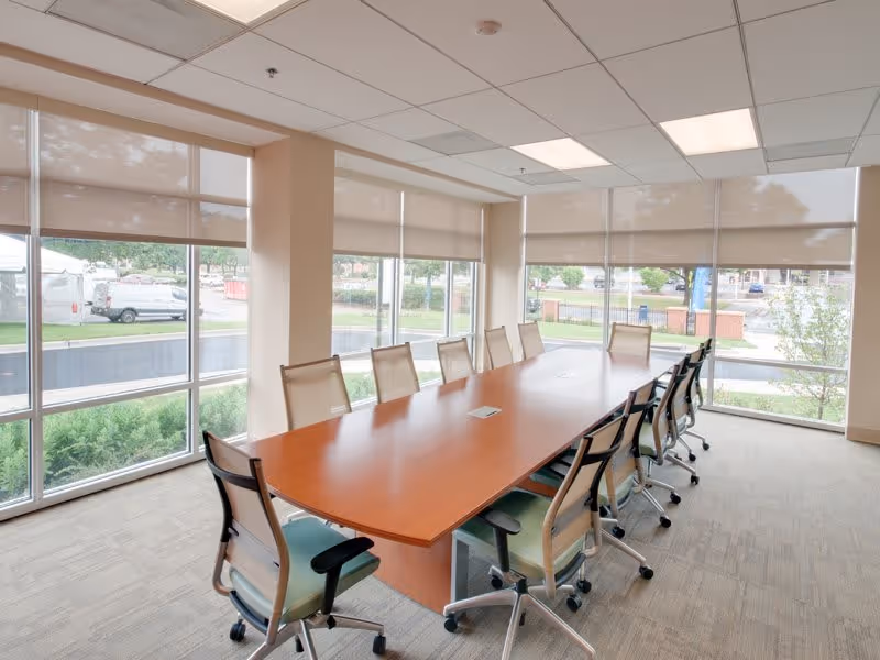 A bright conference room with a long wooden table surrounded by twelve office chairs on wheels. Large windows with beige roller blinds let in natural light and offer a view of greenery and parked vehicles outside.