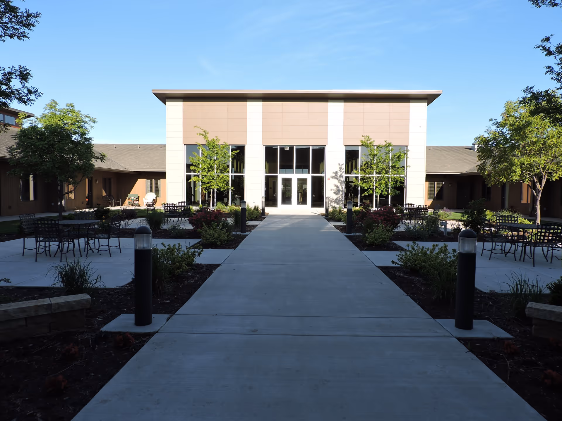 Wide concrete pathway leading to the entrance of a modern building with large glass doors and windows, surrounded by landscaped gardens with small trees, shrubs, and outdoor seating areas with metal tables and chairs under a clear blue sky.