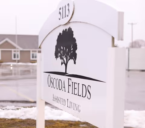 White outdoor sign for Oscoda Fields Assisted Living with a black tree logo and the number 5113 on top, set against a background of a parking lot and buildings on a cloudy day.