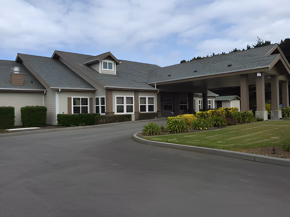Front entrance with covered porte-cochère of a senior living building, driveway, and landscaped shrubs.