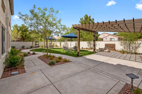 Outdoor patio area at Oakmont of the Lakes featuring a pergola casting shadows on a concrete surface, a small grassy area with a table and chairs under a blue umbrella, young trees, and landscaping with shrubs and mulch, all enclosed by a white wall with neighboring houses visible in the background under a clear blue sky.
