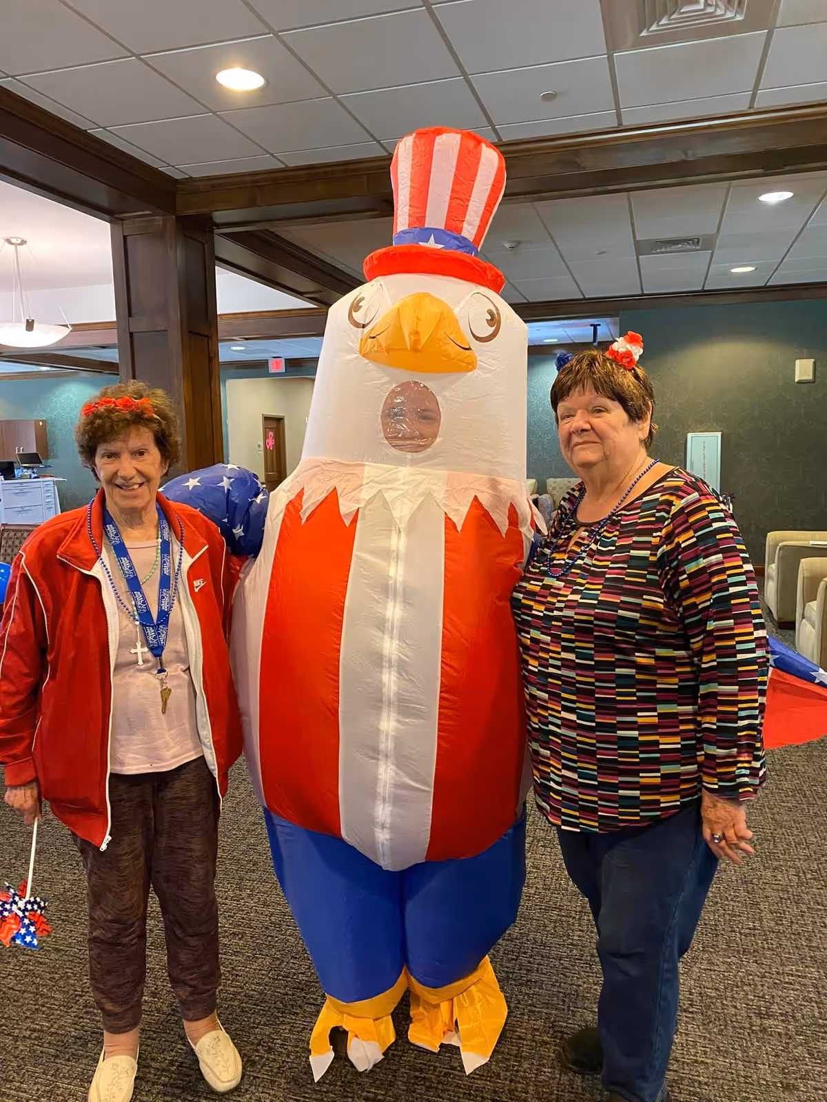Two elderly women standing indoors on either side of a person dressed in a large inflatable patriotic eagle costume with red, white, and blue colors. The setting appears to be a common area or lounge with carpeted floors and wooden beams.
