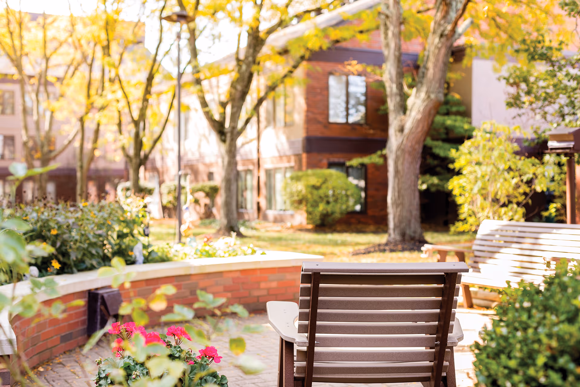 Outdoor seating area in a garden with wooden chairs and benches surrounded by trees and flowering plants, with a building visible in the background under bright sunlight.