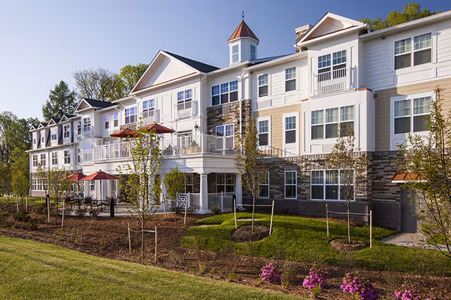 Exterior view of a senior living facility building with three stories, featuring white and beige siding, stone accents, multiple windows, balconies with red umbrellas, and landscaped grounds with young trees and flowers.