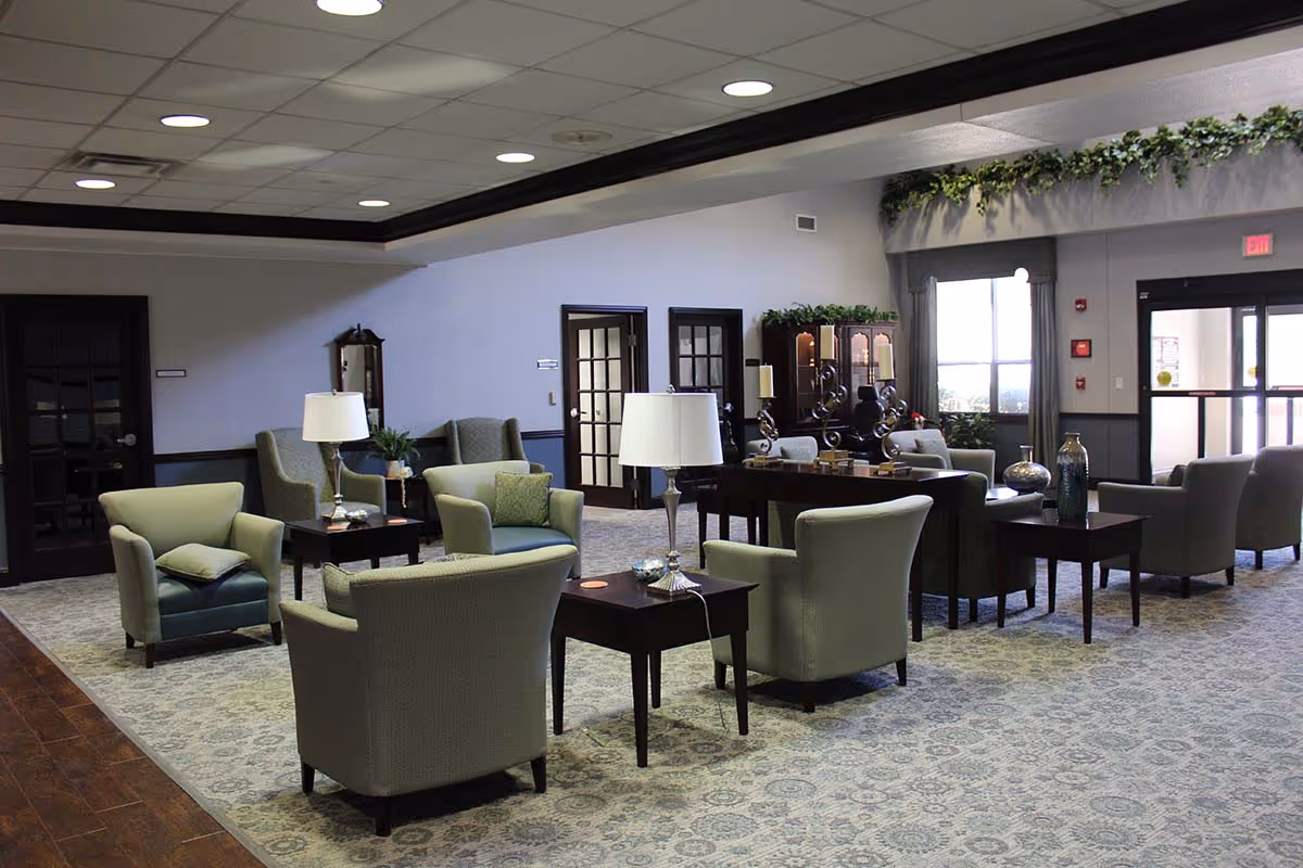 A spacious senior living facility common area with multiple armchairs and side tables arranged on a patterned carpet. The room features ceiling lights, a wooden floor section, decorative plants, and large windows letting in natural light. There are dark-framed glass doors and a cabinet with decorative items in the background.