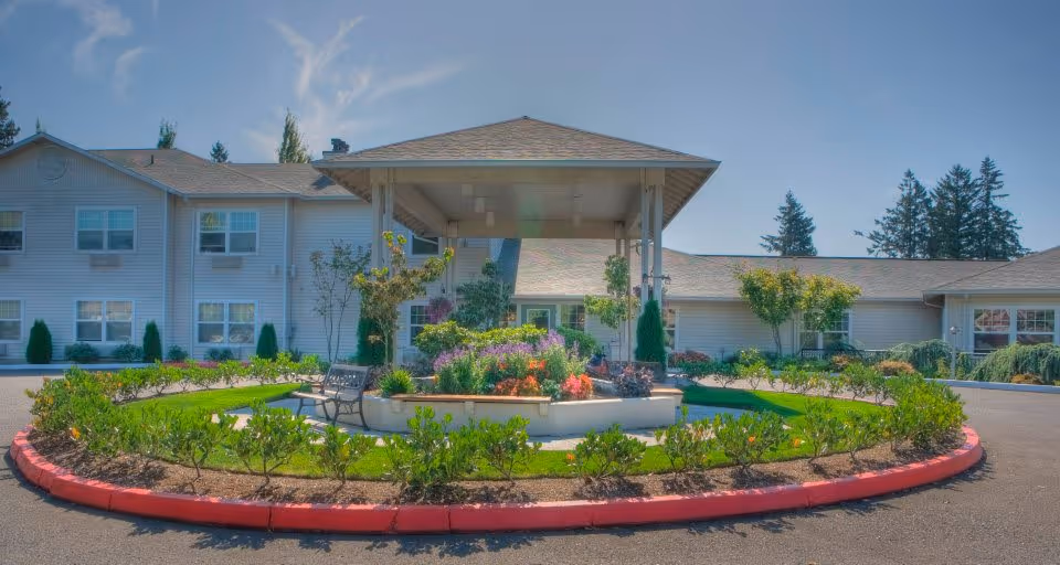 Exterior view of Tanner Spring Assisted Living & Memory Care facility showing a circular driveway with a landscaped garden in the center, featuring various plants and flowers under a covered structure. The building is two stories with multiple windows and surrounded by trees under a clear blue sky.