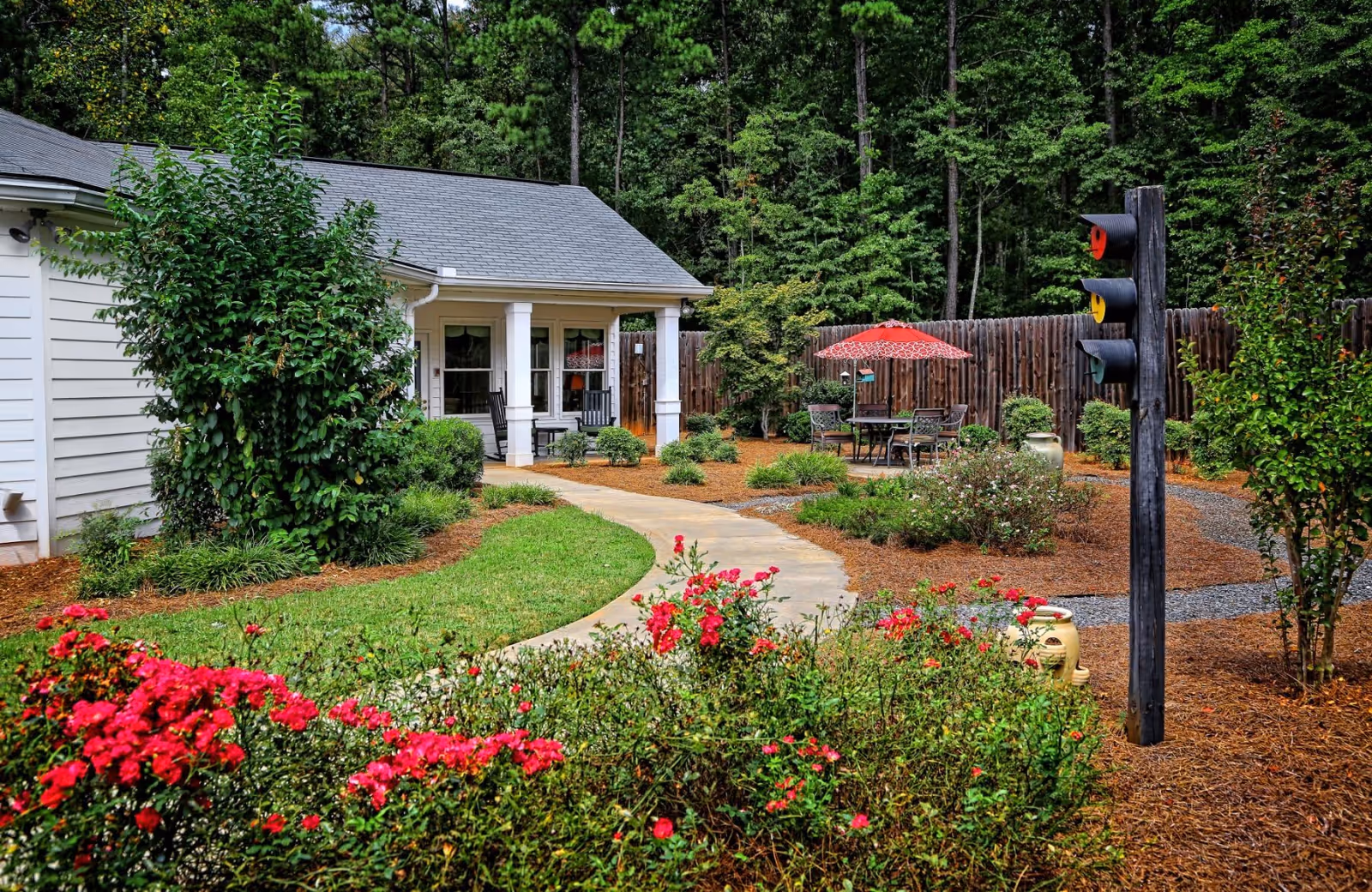 A peaceful outdoor garden area at The Woods Senior Care featuring a curved concrete pathway, vibrant red flowers, green shrubs, and trees. There is a white building with a porch that has two black rocking chairs. In the background, there is a patio table with chairs and a red umbrella, surrounded by a wooden fence and tall trees.