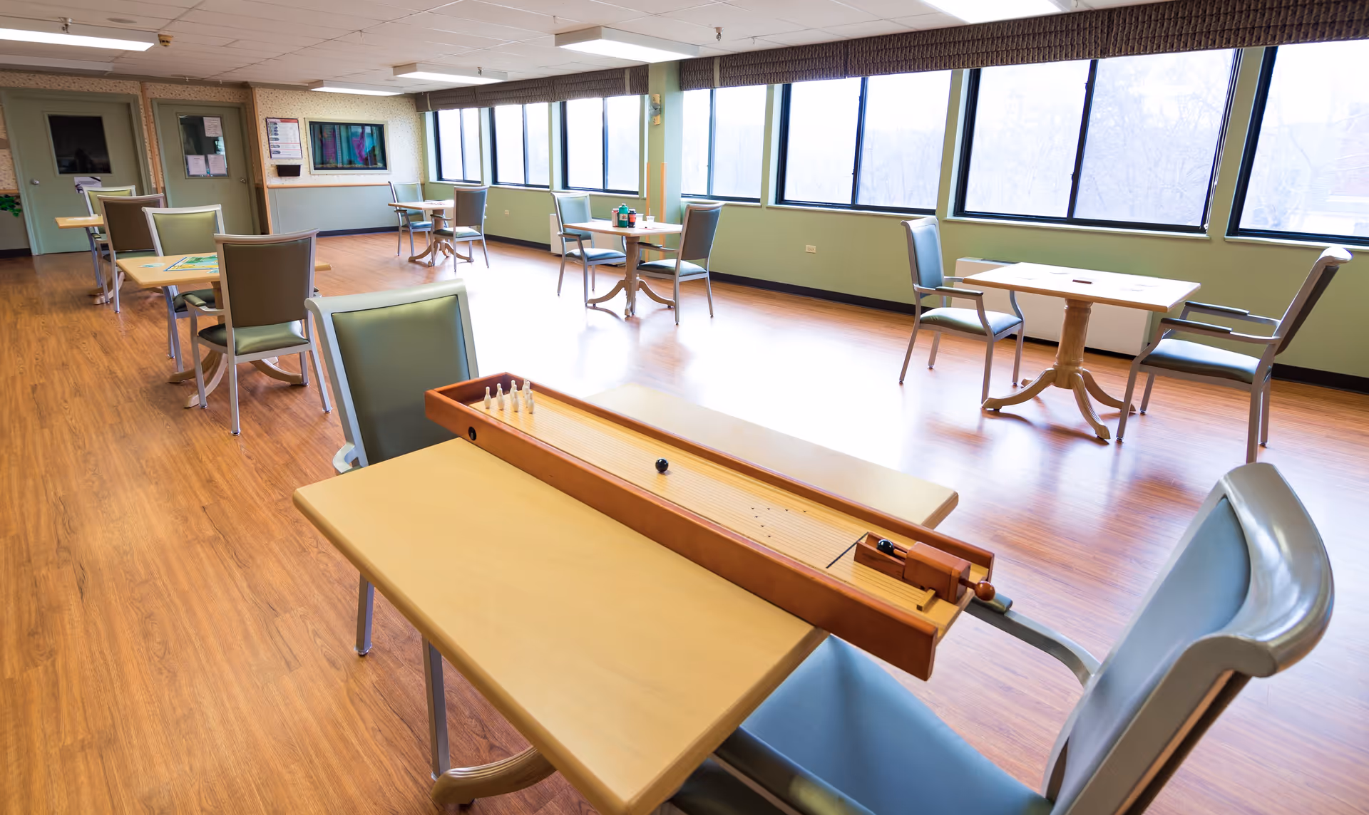 Bright, spacious dining/activity room with tables and chairs and a tabletop shuffleboard game near large windows.