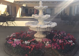 A white multi-tiered fountain surrounded by a circular flower bed filled with pink, red, and white flowers in an outdoor courtyard area with tables and chairs under a covered walkway.