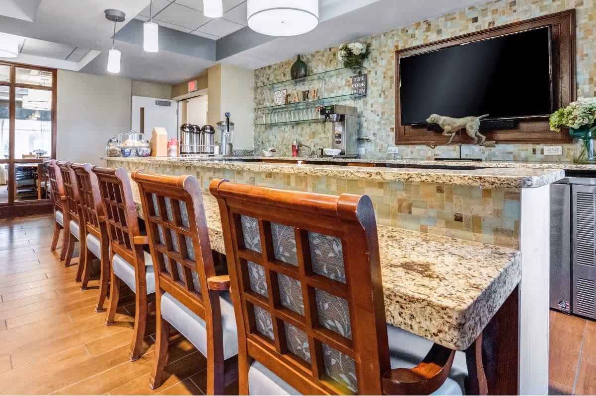 Interior view of a senior living facility's cafe area featuring a long granite countertop with wooden chairs lined up along it. The back wall has a mosaic tile design with shelves holding glassware and decorative items. A large flat-screen TV is mounted on the wall above the counter, and there are pendant lights hanging from the ceiling.