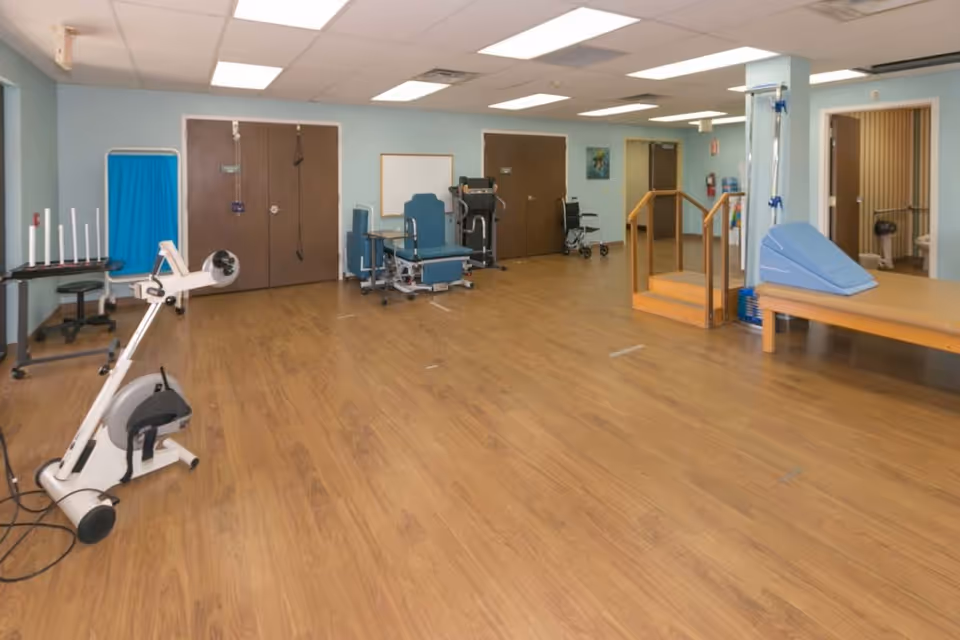 A spacious rehabilitation or physical therapy room with wooden flooring, exercise equipment including a stationary bike, therapy chairs, a set of stairs with handrails, and a padded therapy table with a blue wedge cushion. The room has light blue walls, a drop ceiling with fluorescent lights, and several doors leading to other areas.