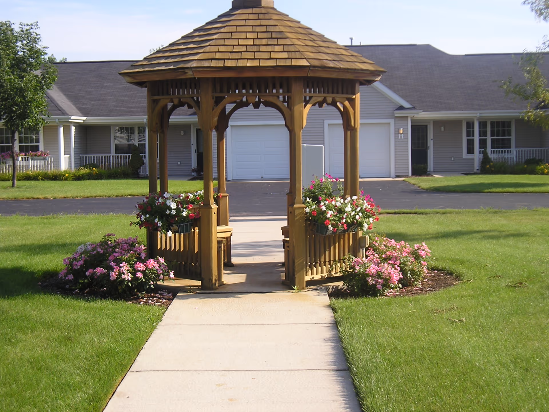 A wooden gazebo with a shingled roof is centered on a concrete pathway surrounded by green grass and flower beds with pink and white flowers. In the background, there are single-story residential buildings with garages and porches.