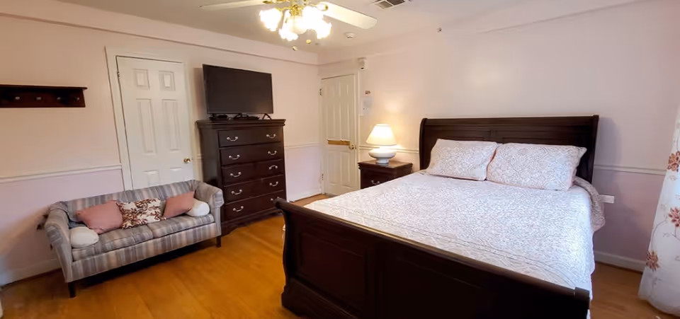 A bedroom with a large dark wooden bed with white and pink patterned bedding, a matching dark wooden dresser with a flat-screen TV on top, a small striped couch with several pillows, a nightstand with a lamp, and two closed doors. The walls are painted light pink with white trim, and there is a ceiling fan with lights above.