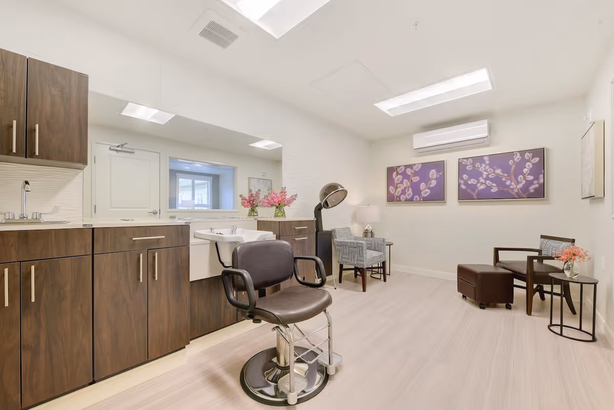 A bright and clean salon room with a black salon chair in front of a white sink and dark wood cabinets. The room has light wood flooring, two armchairs with a small table and lamp between them, and two purple floral paintings on the wall. There is an air conditioning unit mounted above the paintings.