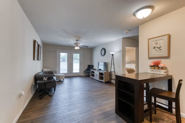A spacious living room area with wood flooring, featuring a small dining table with two chairs on the right, a TV on a wooden stand against the far wall, a floor lamp, and two armchairs near glass double doors leading outside. A doorway to a bedroom is visible on the right side of the room.