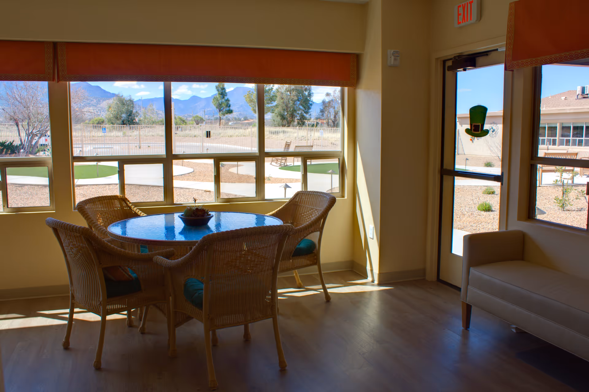 A bright interior room with a round glass-top table surrounded by four wicker chairs with cushions. Large windows provide a view of an outdoor garden area with pathways, trees, and mountains in the background. There is a beige couch near a glass door with an exit sign above it.