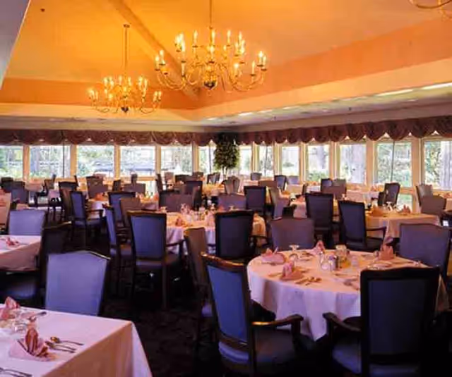 A spacious dining room with multiple round tables covered with white tablecloths and set with pink napkins, silverware, and glassware. The room features large windows with valance curtains, elegant chandeliers hanging from the ceiling, and dark upholstered chairs around each table.