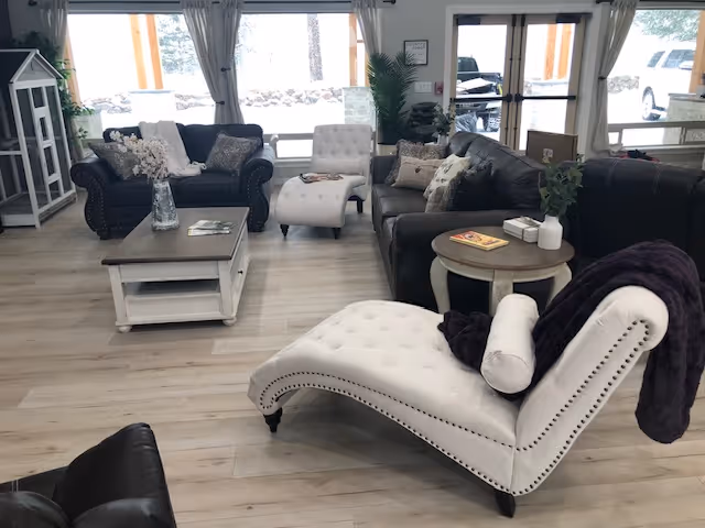 A bright and cozy living room with light wood flooring, featuring a white tufted chaise lounge with a purple throw, dark leather sofas, a white coffee table with a vase of flowers, and large windows with curtains letting in natural light.