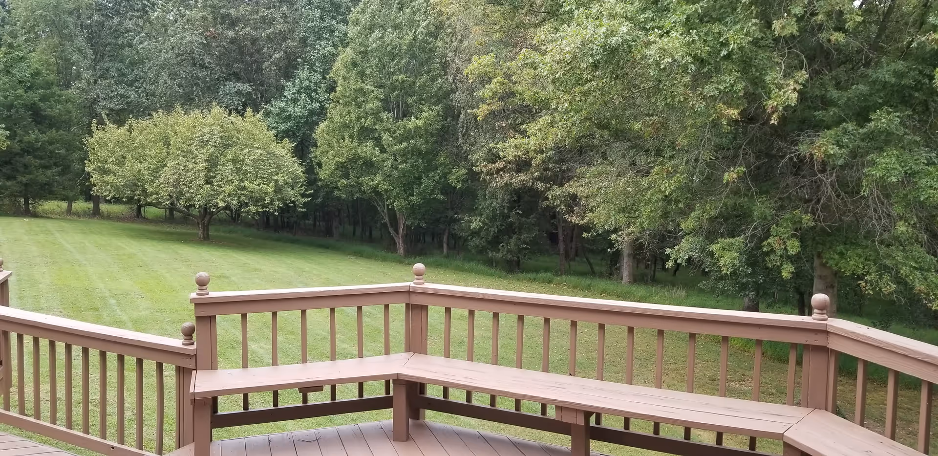 View from a wooden deck with built-in bench seating overlooking a well-maintained grassy yard and a backdrop of dense green trees.