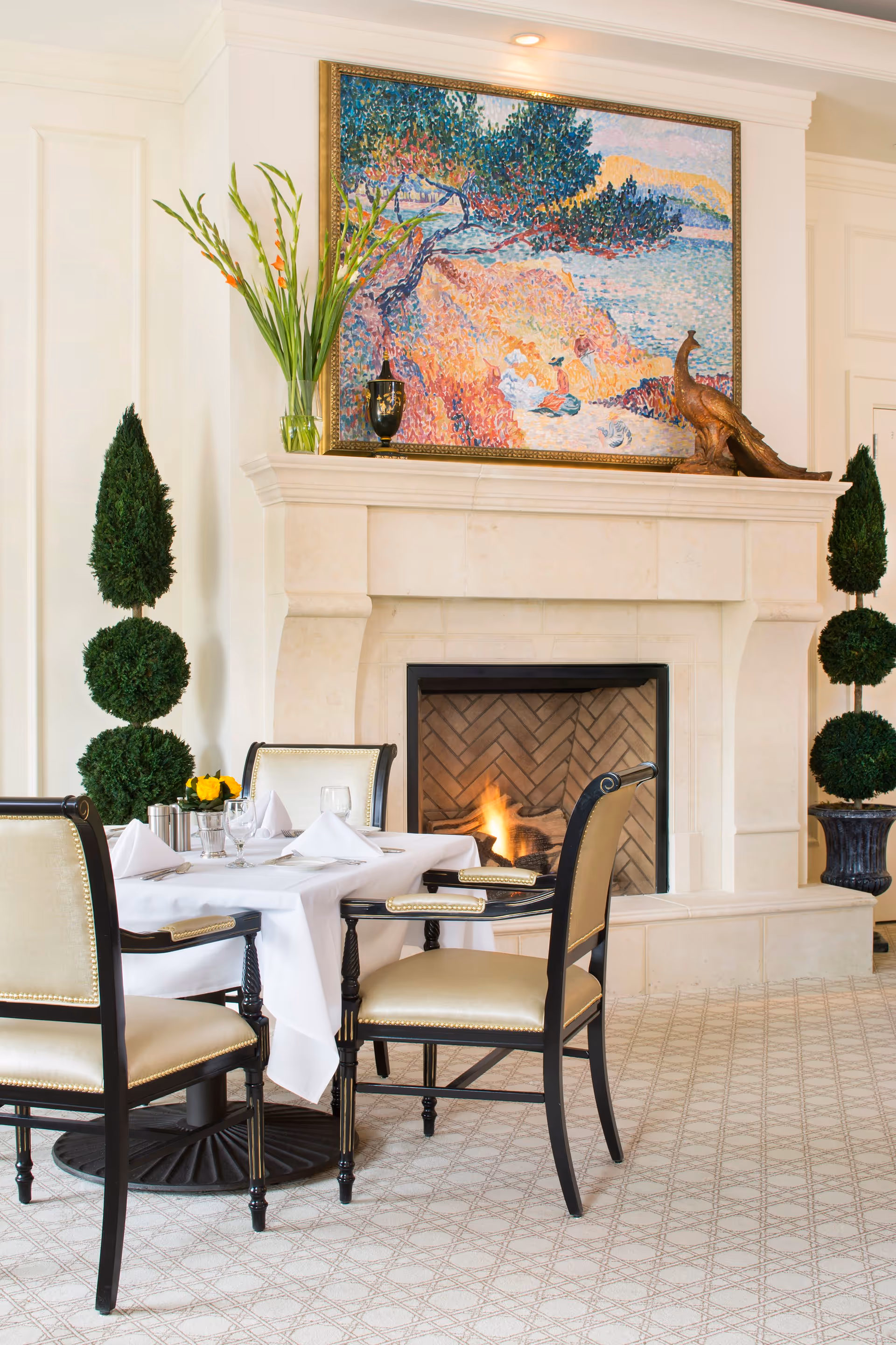 A cozy dining area with a table set for four, featuring white tablecloth and napkins, elegant chairs with beige upholstery, a lit fireplace with a stone mantel, a colorful painting above the fireplace, and decorative topiary plants on either side.