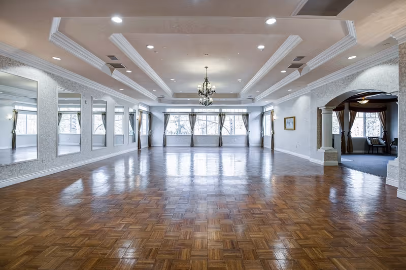 A spacious, empty room with polished wooden parquet flooring, large windows with curtains allowing natural light, decorative ceiling with recessed lighting and chandeliers, and mirrors on one wall. An adjacent room is visible through an arched opening with columns.
