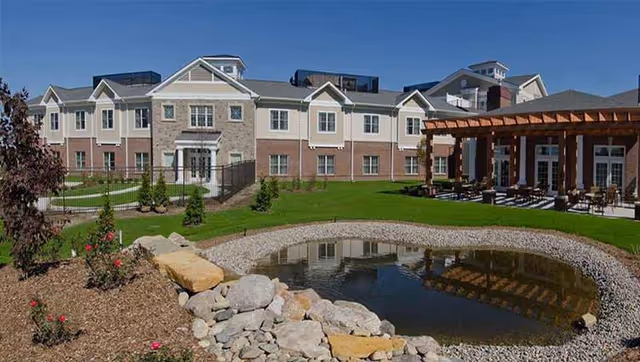 Exterior view of St Clare Commons senior living facility showing a large two-story building with multiple windows, a well-maintained lawn, a small pond surrounded by rocks, and a wooden pergola with outdoor seating on a sunny day.