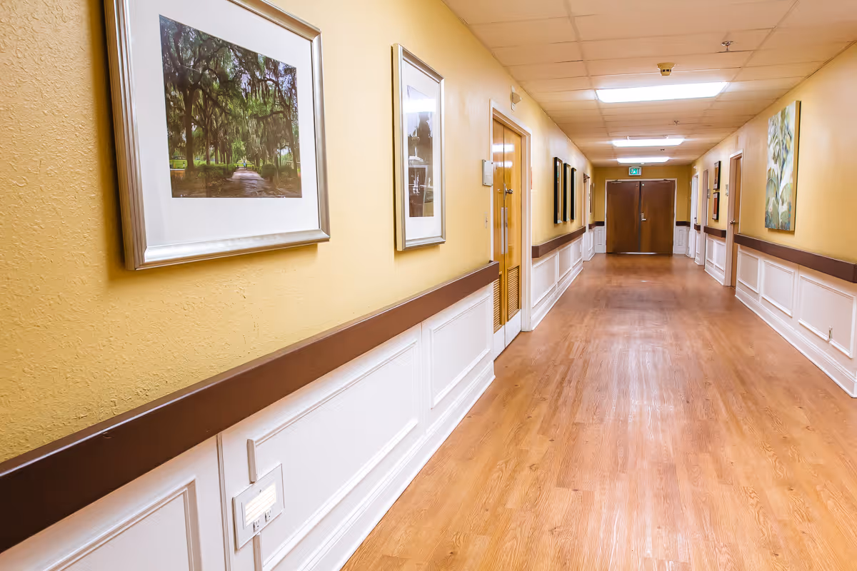 A long, well-lit hallway with wooden flooring and yellow walls. The walls are decorated with framed pictures and paintings. There are several doors along the hallway and a double door at the end.