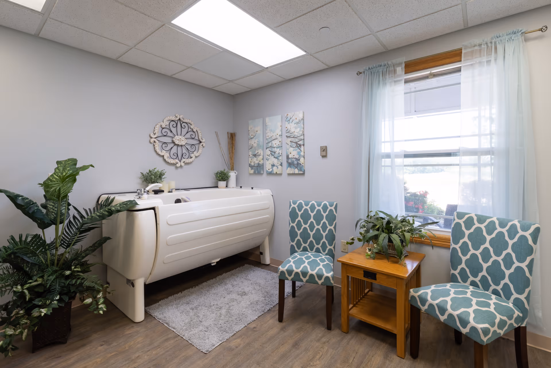 A bright interior bathing room with a white walk-in tub, two patterned chairs, plants, and a window with sheer curtains.