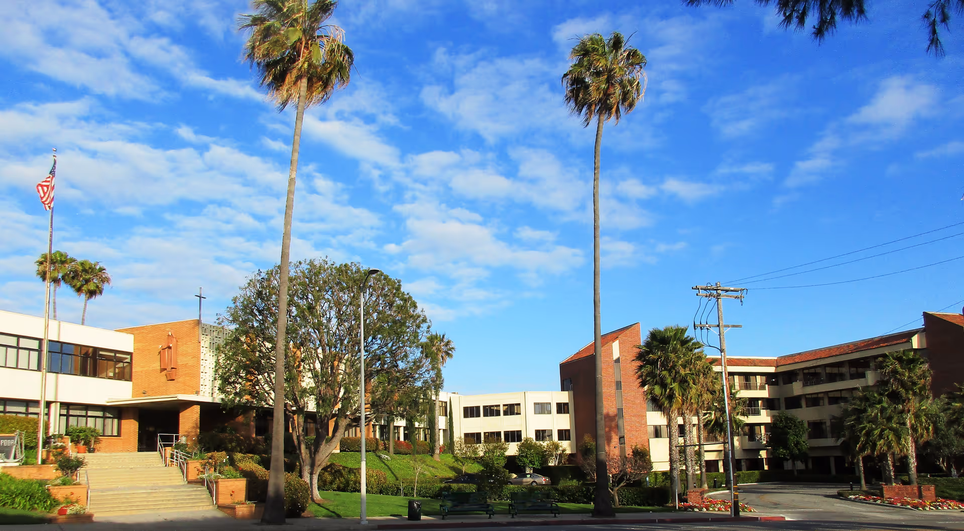 Exterior view of a senior living facility with multiple buildings, palm trees, a flagpole with an American flag, and a clear blue sky with some clouds.