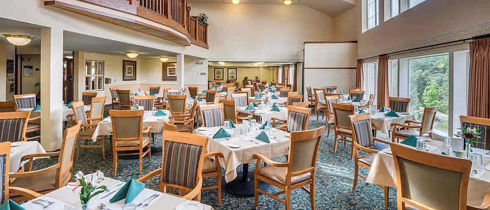 A spacious dining room with multiple tables covered in white tablecloths, each set with green folded napkins, plates, glasses, and silverware. Wooden chairs with striped cushions surround the tables. Large windows on the right side let in natural light and offer a view of greenery outside. The room features a carpeted floor with a floral pattern and a high ceiling with a balcony area above.