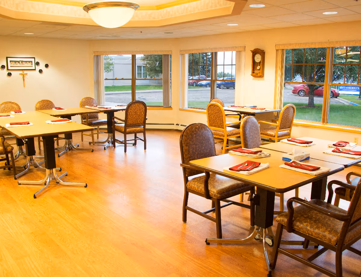 A bright dining room with several tables and chairs arranged neatly. Each table is set with red napkins, silverware, and white placemats. Large windows provide a view of the outside parking area and greenery. The room has wooden flooring, a ceiling light fixture, a wall clock, and a religious cross with a framed picture on the wall.