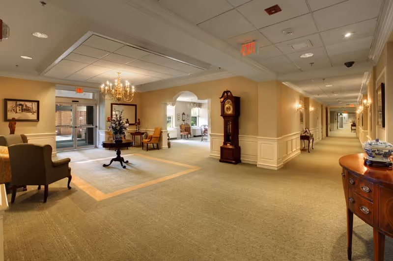 A spacious, well-lit hallway in a senior living facility with beige walls, carpeted floors, and classic wooden furniture including a grandfather clock, chairs, and tables. A chandelier hangs from the ceiling, and there are exit signs and framed artwork on the walls. The hallway leads to other rooms and an entrance with glass doors.
