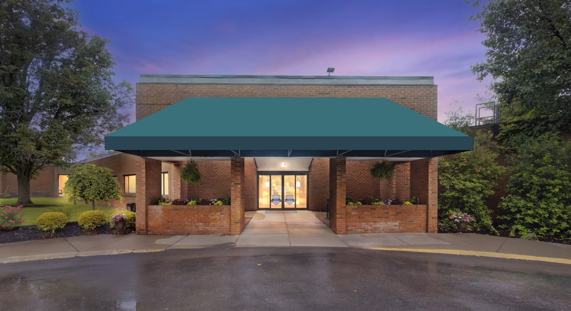 Entrance of a brick building with a green awning over the walkway, flanked by brick planters with hanging plants and flowers, surrounded by trees and shrubs under a purple evening sky.