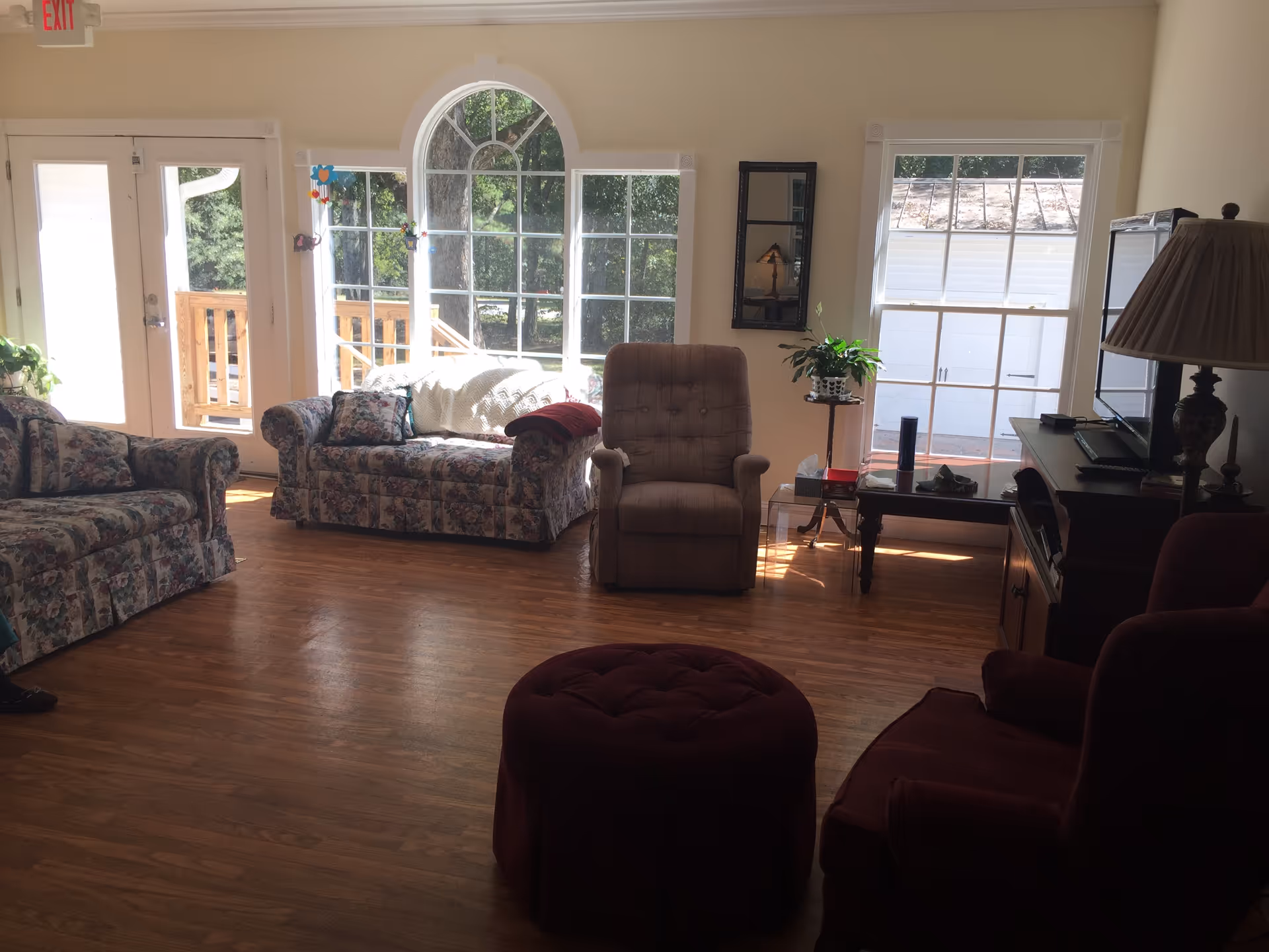 A cozy living room with floral patterned sofas, a beige recliner, a maroon armchair, and a matching ottoman. The room has large windows letting in natural light, a wooden floor, a TV on a stand, a side table with a plant, and a lamp. There is a door with an exit sign above it leading outside to a wooden deck.