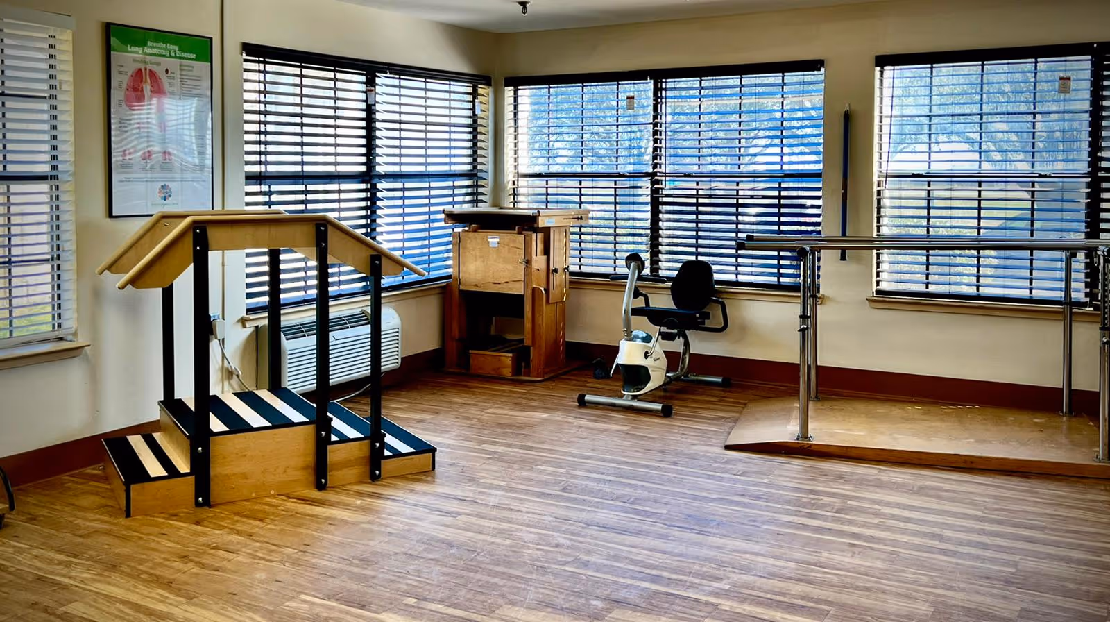 A rehabilitation therapy room with wooden stairs equipped with handrails, a stationary exercise bike, a wooden therapy cabinet, and parallel bars for walking exercises. The room has large windows with horizontal blinds allowing natural light to enter, and a wooden floor.