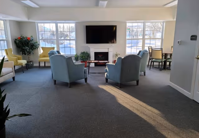 Sunlit communal living room with armchairs arranged around a fireplace and wall-mounted TV in front of large windows.