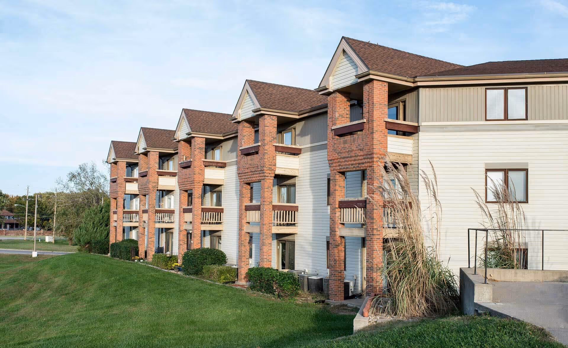 Exterior view of a multi-story senior living facility building with brick and siding facade, balconies, and a grassy lawn in front under a partly cloudy sky.