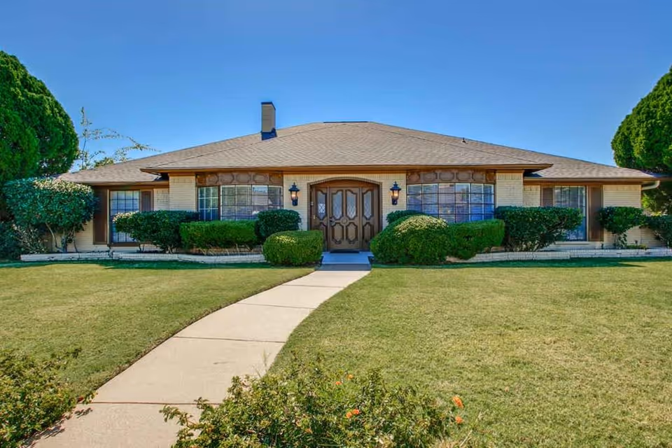 Front exterior view of a single-story building with a large, well-maintained lawn and a concrete walkway leading to a double wooden door entrance. The building has large windows with decorative trim and is surrounded by neatly trimmed bushes and trees under a clear blue sky.