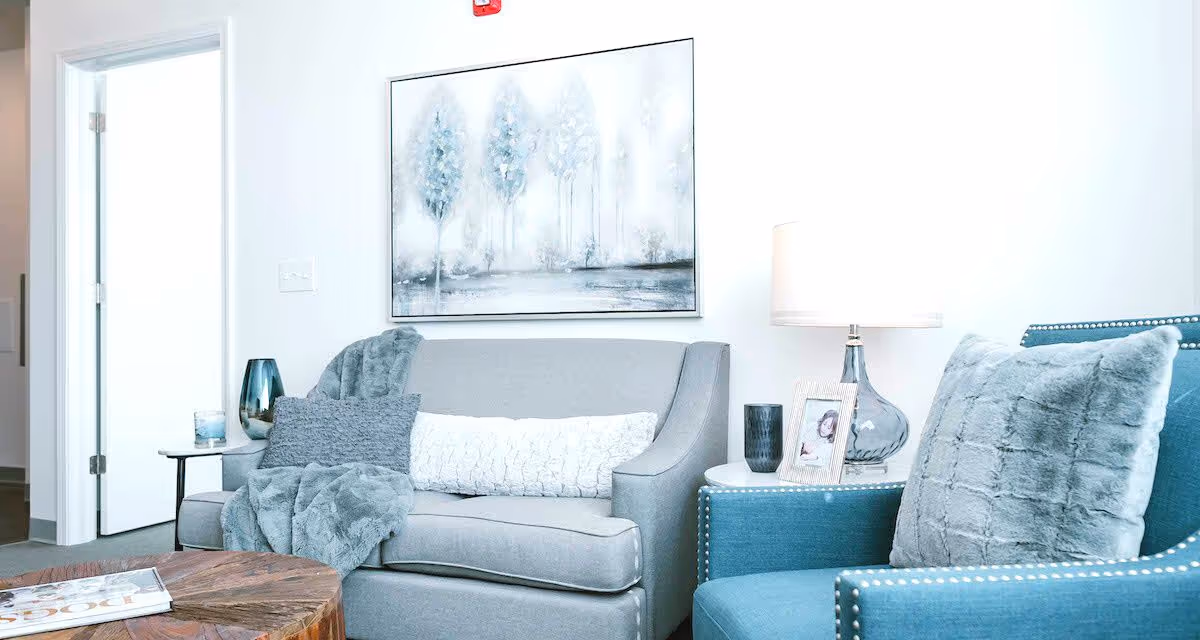 A cozy living room area with a gray loveseat adorned with a gray throw blanket and pillows, a blue armchair with a gray pillow, a round wooden coffee table with a magazine on it, a white side table with a lamp, a framed photo, and decorative items. A painting of trees hangs on the white wall above the loveseat.