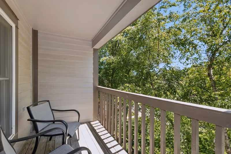 A small balcony with two black metal chairs with mesh seats and backs, overlooking green leafy trees under a clear blue sky.