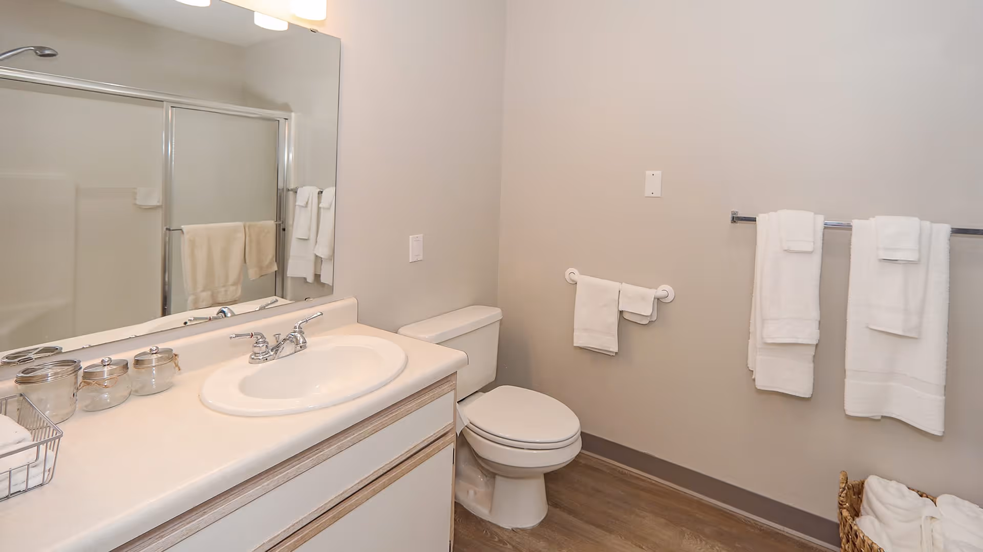 A clean bathroom with a white sink and countertop, a large mirror above the sink, a toilet, and a shower with sliding glass doors. Several white towels are hanging on towel racks and a basket with additional towels is on the floor.