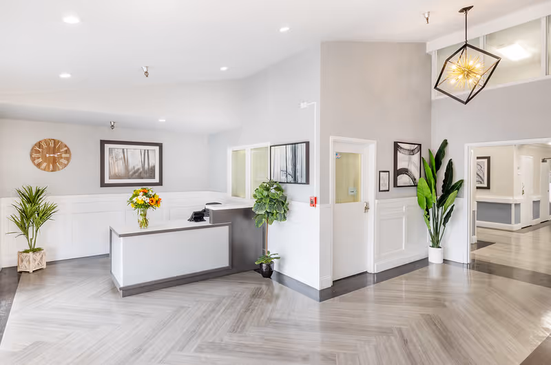 A bright and modern reception area in a nursing and rehabilitation center with a white reception desk adorned with a vase of flowers. The space features light gray walls with white wainscoting, a wooden clock, framed artwork, and several green potted plants. The floor has a light wood pattern, and a geometric pendant light hangs from the ceiling.